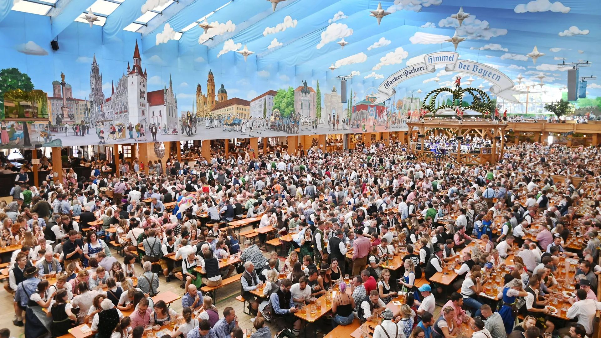 Tenda lotada na Oktoberfest de Munique, com o teto pintado de céu e mesas compridas cheias de gente.