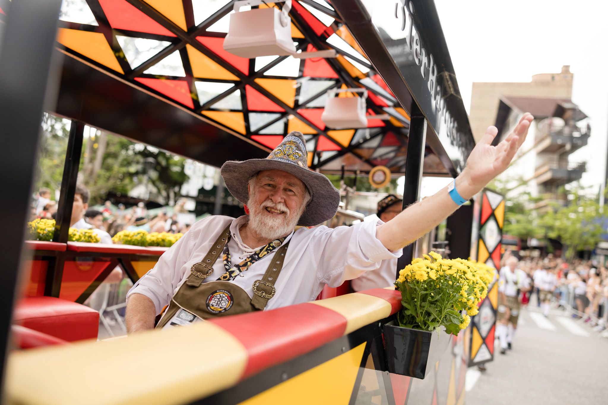 Senhor de chapéu típico sorri e acena de um carro alegórico com vitral colorido, no desfile da Oktoberfest de Blumenau de 2024.
