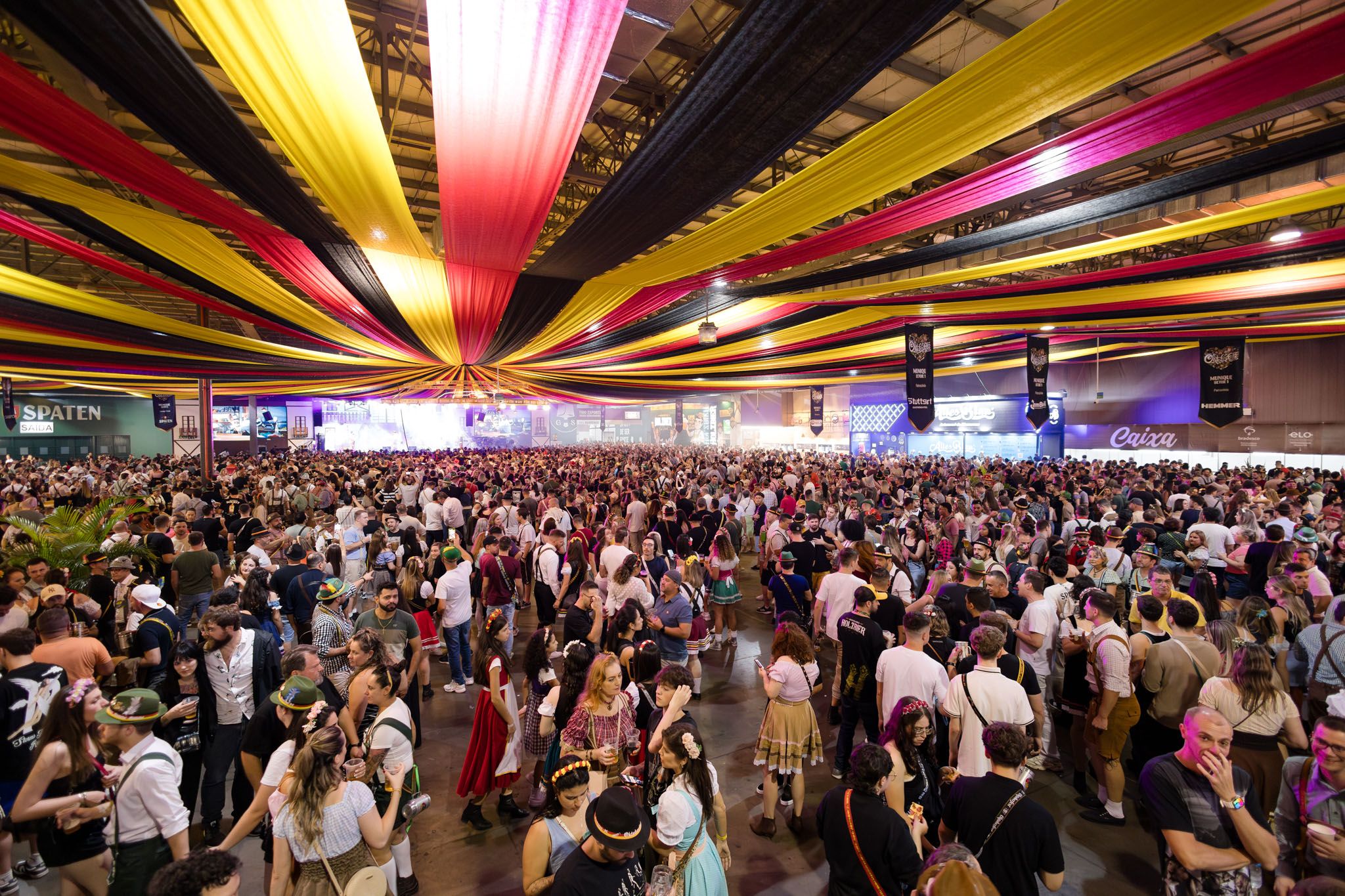 Pavilhão da Oktoberfest de Blumenau de 2024 lotado, visto do alto, com faixas amarelas, vermelhas e pretas no teto.