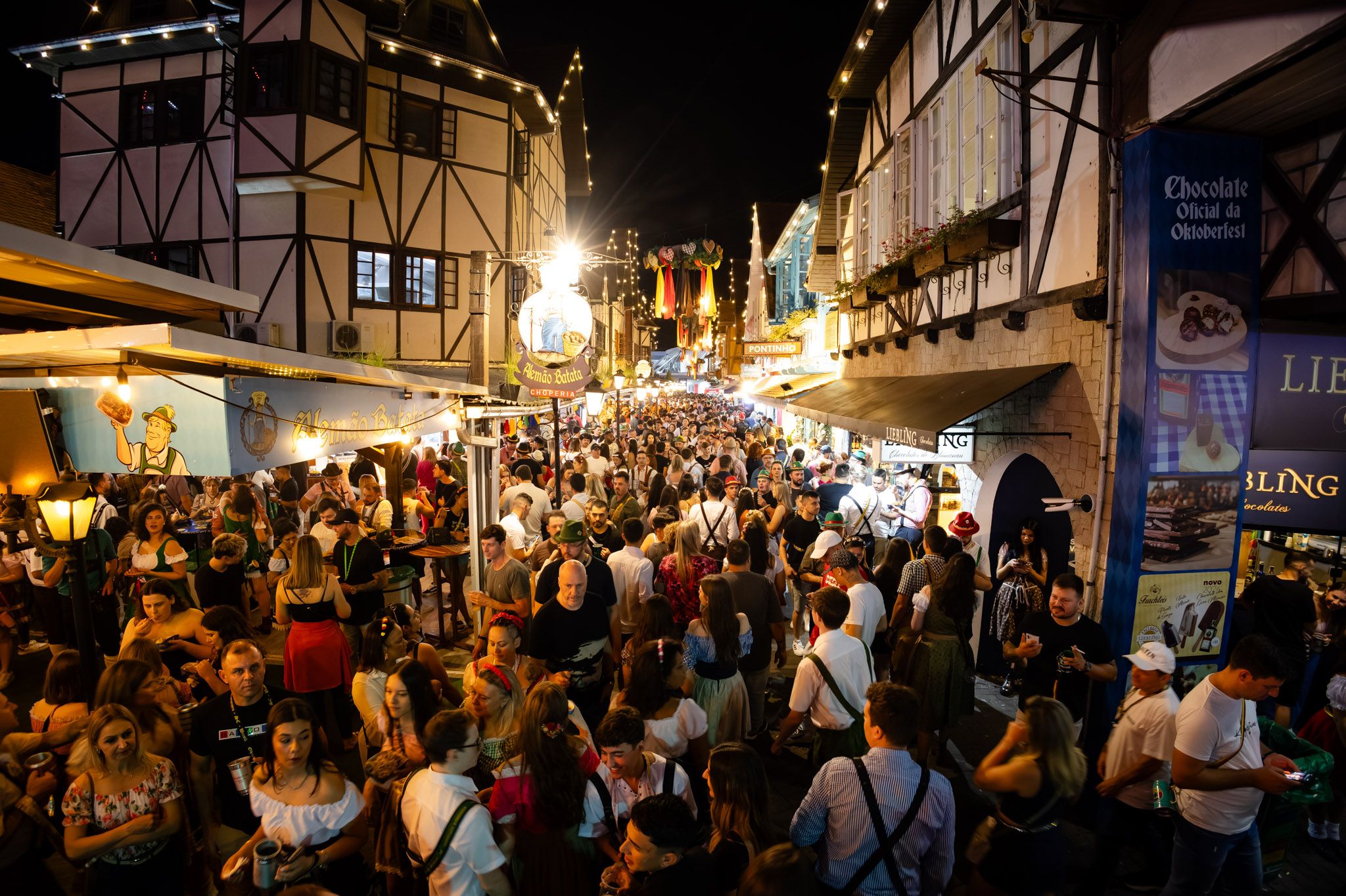 Rua da Vila Germânica à noite, com casas em estilo enxaimel iluminadas e o público da Oktoberfest de Blumenau de 2024.